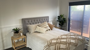 Un dormitorio con un cabecero gris con textura, un ventanal con persianas, una cama con colcha color crema y una mesita de noche con una planta y adornos.