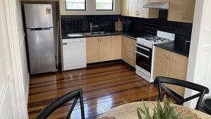 Una cocina funcional con refrigerador de acero inoxidable, lavavajillas, estufa y armarios de madera. Dos ventanas proporcionan luz natural.