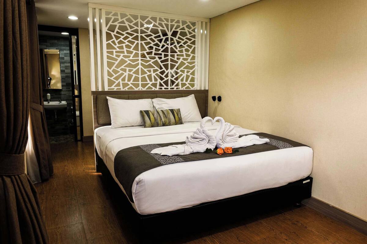 A hotel room with a large bed, a decorative wall panel, a bathroom visible through an open door, and a wooden floor.