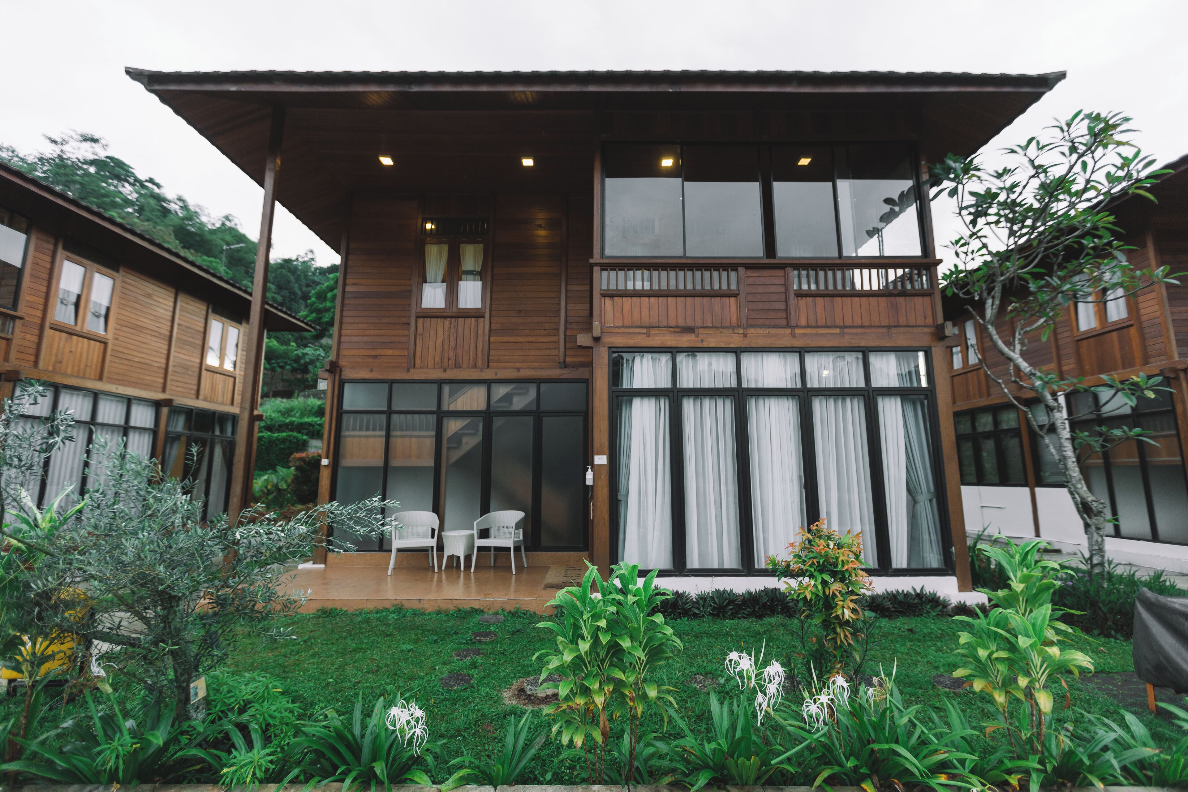 A two-story wooden house with a balcony, a covered patio, and a garden with various plants.