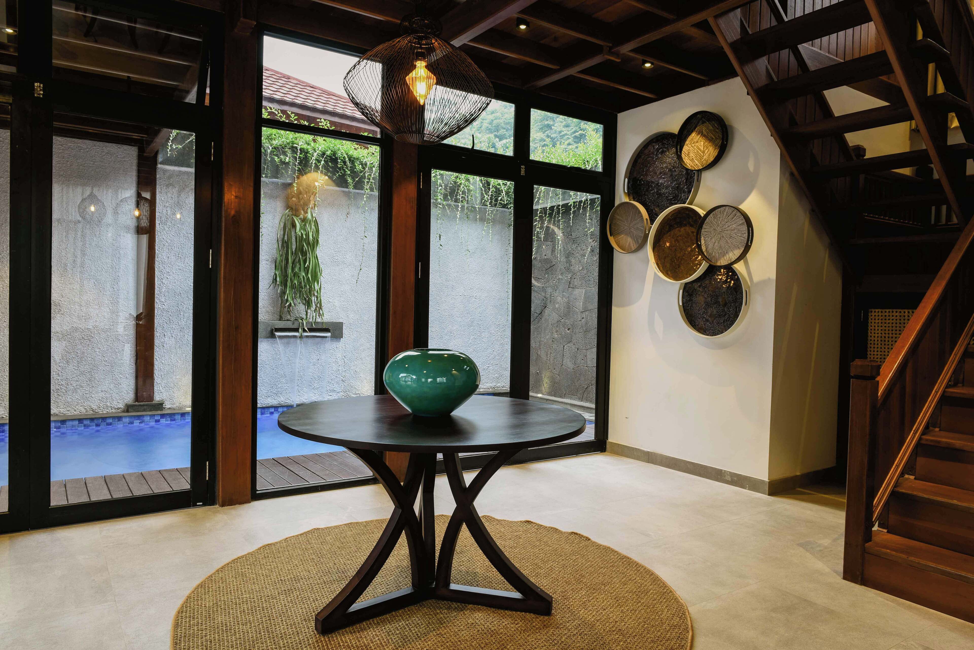A modern interior with a round table, a hanging light fixture, and a view of an outdoor pool through glass doors.