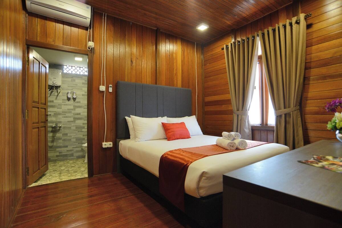 A hotel room with wooden paneling, a bed with white and red bedding, a bedside table with towels, and a window with curtains.