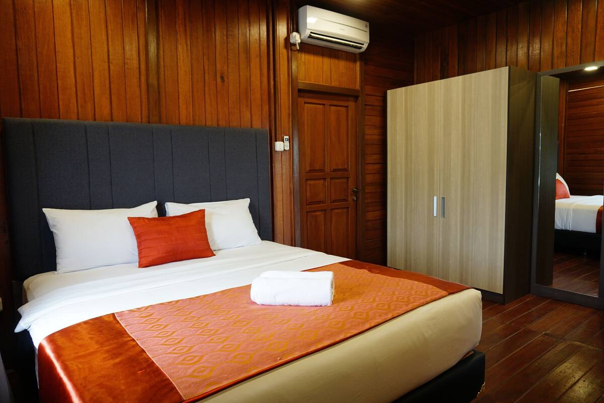 A hotel room with a large bed, wooden walls, a wardrobe, and an air conditioning unit.