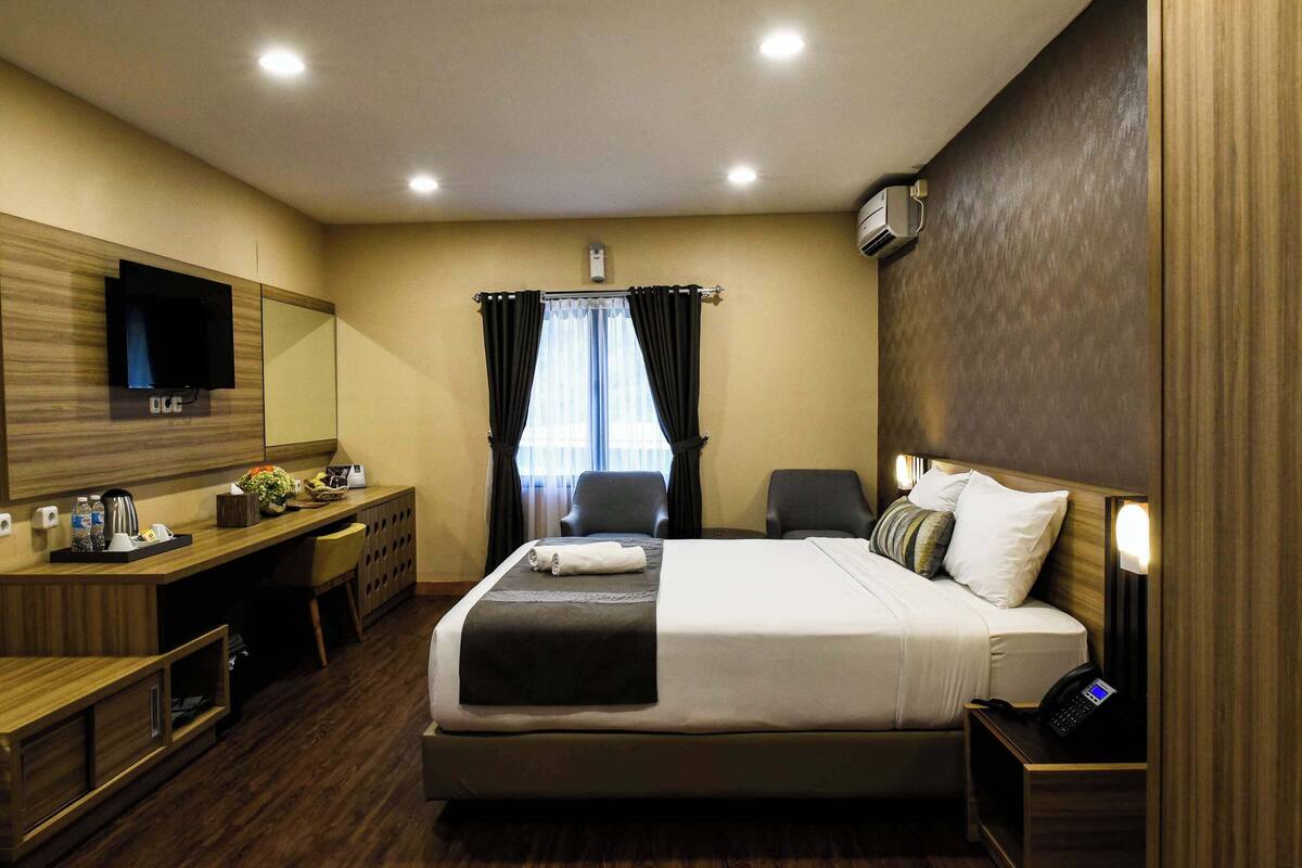 A hotel room with a large bed, a desk with a computer, a TV, and a window with curtains.