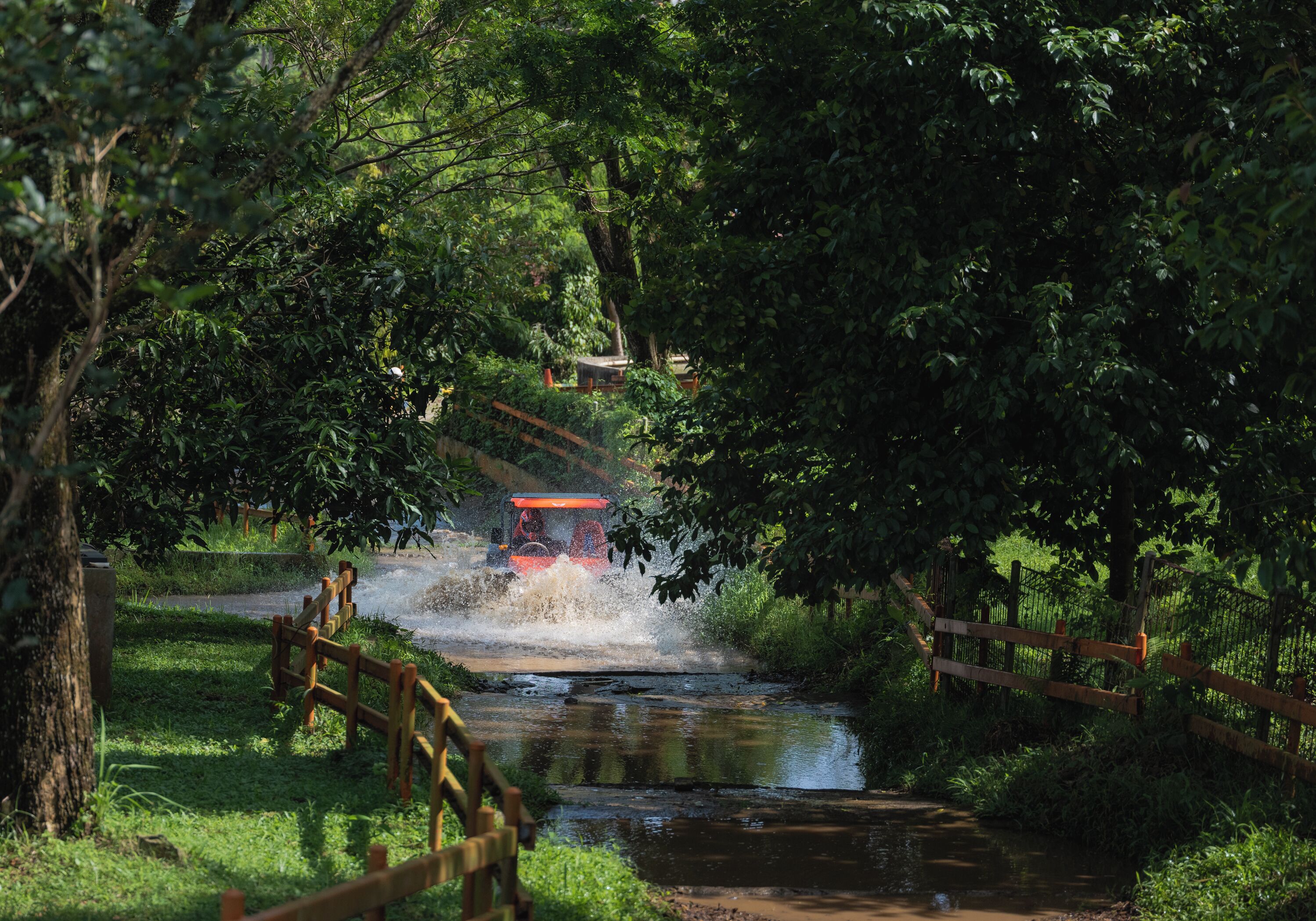A red vehicle crossing a shallow water crossing in a lush, green area with wooden fences and trees.