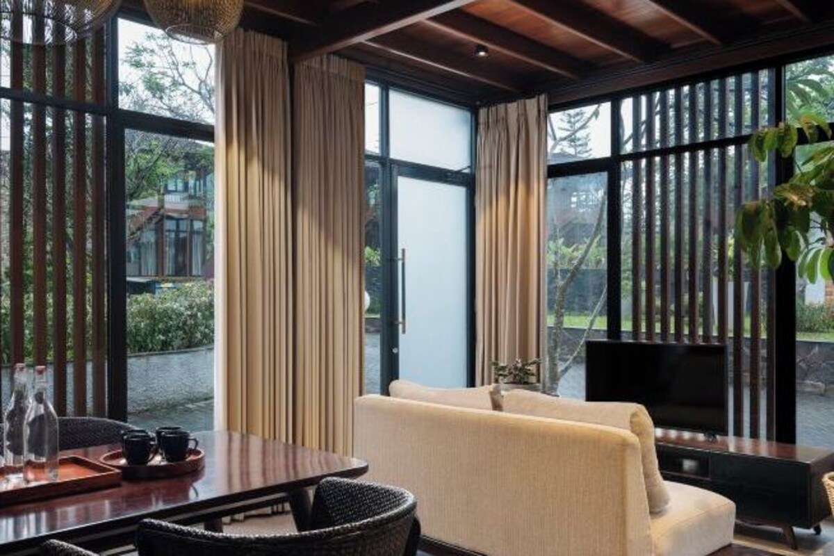 A modern living room with a sofa, a wooden coffee table, and a television. Large windows with vertical blinds allow natural light and views of greenery outside.
