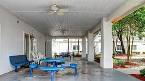 Un espace extérieur abrité, avec des tables et des chaises, un ventilateur de plafond et une vue sur un bâtiment et des arbres.