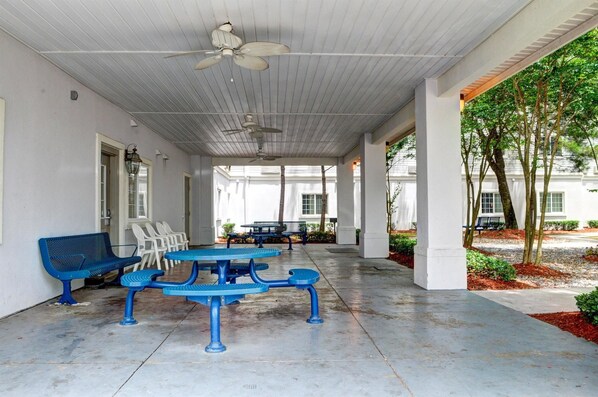 A covered outdoor seating area with tables and chairs, a ceiling fan, and a view of a building and trees. - Studio 6 New Orleans (New Orleans)