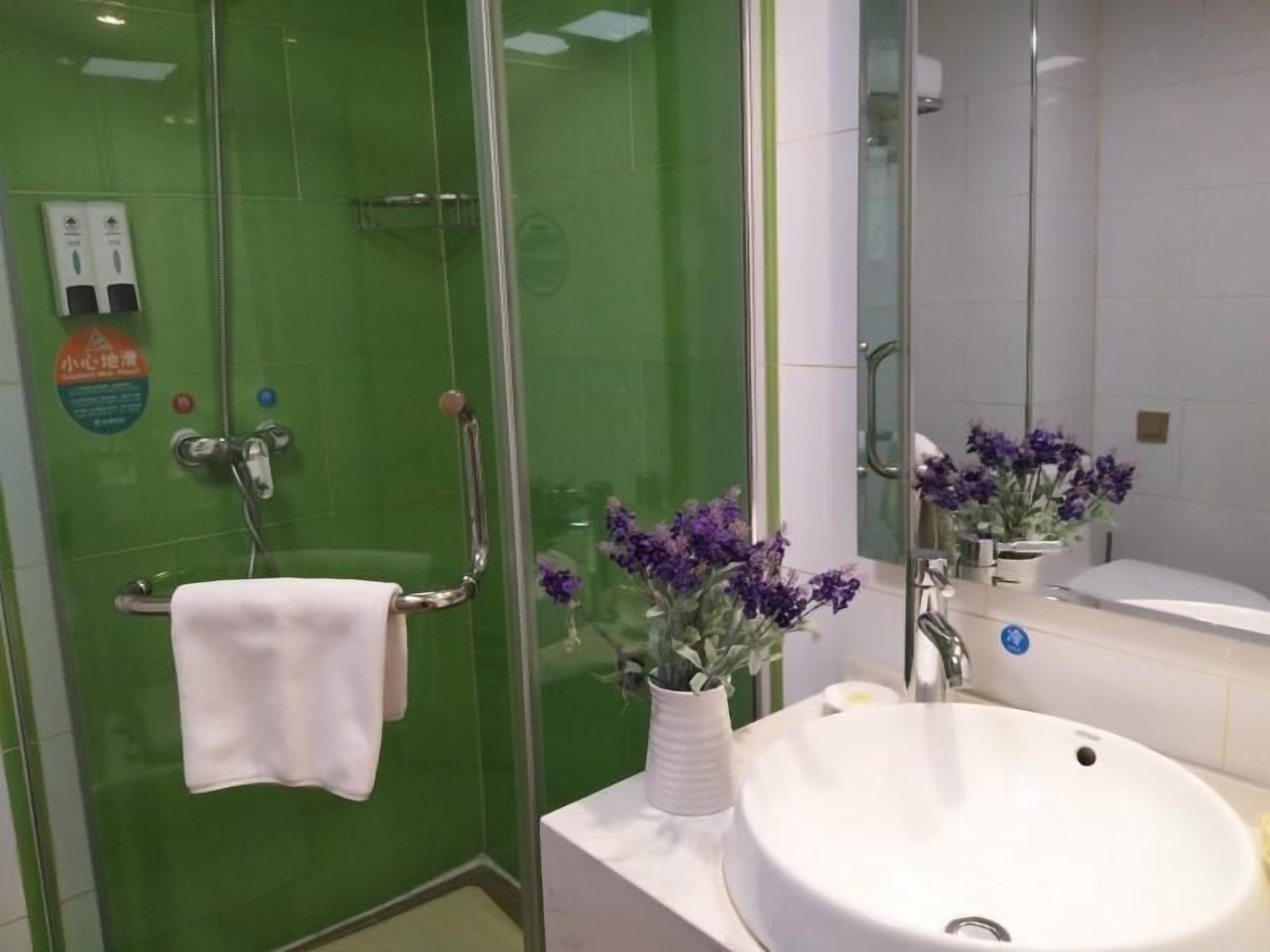A bathroom with a green-tiled shower, a white sink, and a mirror reflecting a purple flower arrangement.