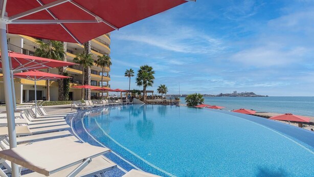 A pool area with lounge chairs and umbrellas, a clear view of the sea, and a building with balconies. - Sonoran Sky Resort 310 Vacationer's Dream Getaway Ocean Front Condo (Puerto Peñasco)