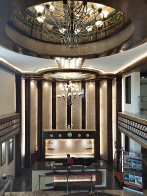 Zona de recepción de un hotel con una imponente lámpara de araña, un indicador de hora y un mostrador de recepción.