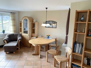 A cozy living room with a round wooden table, four chairs, a grey armchair, a bookshelf, and a window with curtains.