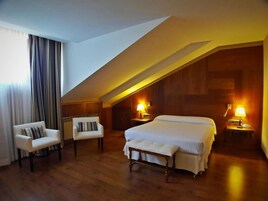 Quarto de hotel com teto inclinado, uma cama grande, duas mesas de cabeceira com luminárias, um banco e uma cadeira.