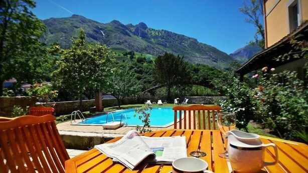 A wooden table with a newspaper, a cup, and a saucer, overlooking a swimming pool with mountains in the background. - Picos de Europa (Cabrales)