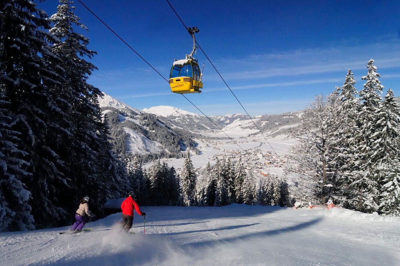 Eine Sesselbahn mit einer Gondel, schneebedeckte Bäume und ein Skifahrer in einer roten Jacke.