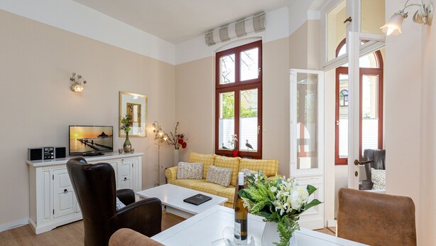 Un salon bien éclairé, avec un canapé, une table basse et des fauteuils. On y trouve également un téléviseur, un vase avec des fleurs et une fenêtre équipée de stores.