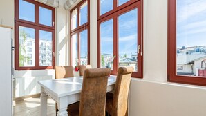 A dining area with a white table and brown chairs, large windows with red frames, and a view of buildings outside.