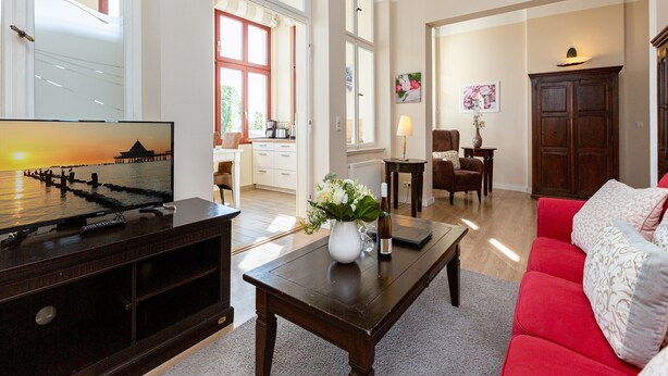 A spacious living room with a red sofa, a coffee table, and a television. There are large windows providing natural light and a view of the outdoors.