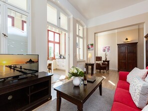 Un salon spacieux avec un canapé rouge, une table basse et un téléviseur. De grandes fenêtres laissent entrer la lumière naturelle et offrent une vue sur l’extérieur.