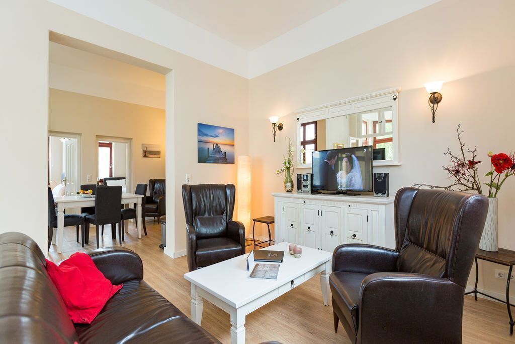 Una sala de estar moderna con una mesa de centro blanca, sofás de cuero, un comedor con una mesa y sillas blancas, y una cocina con un televisor y armarios.