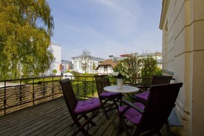Een balkon met paarse, beklede stoelen en een kleine ronde tafel, met uitzicht op een straat met gebouwen en bomen.