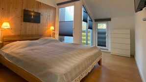 Une chambre à coucher avec une tête de lit en bois, un lit recouvert d’une couvre-lit à motifs, deux lampes de chevet, un téléviseur fixé au mur et une porte-fenêtre coulissante donnant sur un balcon.