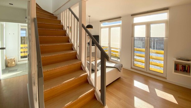 Un intérieur moderne avec un escalier en bois, un canapé blanc et de grandes fenêtres donnant sur une rambarde jaune à l’extérieur.