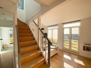 Interior moderno com escada de madeira, sofá branco e janelas amplas com vista para um corrimão amarelo lá fora.