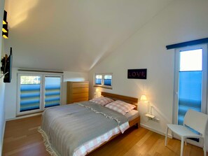 Quarto com cama, duas luminárias de cabeceira, uma cadeira, janela com persianas e um aviso fixado na parede.