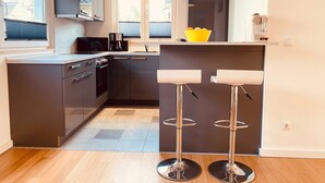Une cuisine moderne avec des meubles de couleur sombre, un four à micro-ondes et une machine à café. On y trouve deux tabourets de bar avec des pieds en métal et des assises blanches.