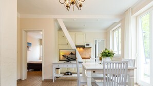 A modern kitchen with white cabinetry, a dining table with chairs, a TV mounted above the table, and a view of the outdoors through large windows. - Villa Amelia, App. 12 (Heringsdorf)