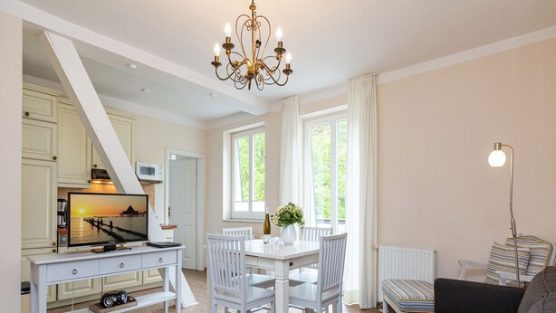 A modern living room with a dining area, white furniture, a chandelier, and a view of greenery outside. - Villa Amelia, App. 12 (Heringsdorf)