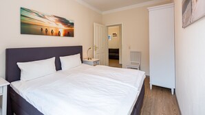 A bedroom with a large bed, a nightstand, a desk, and a chair. There is a painting on the wall and a door leading to another room. - Villa Amelia, App. 12 (Heringsdorf)
