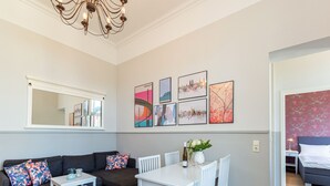 Un salon moderne avec un canapé gris foncé, une table à manger blanche et des chaises. On y trouve un lustre, un miroir et plusieurs œuvres d’art encadrées accrochées au mur.