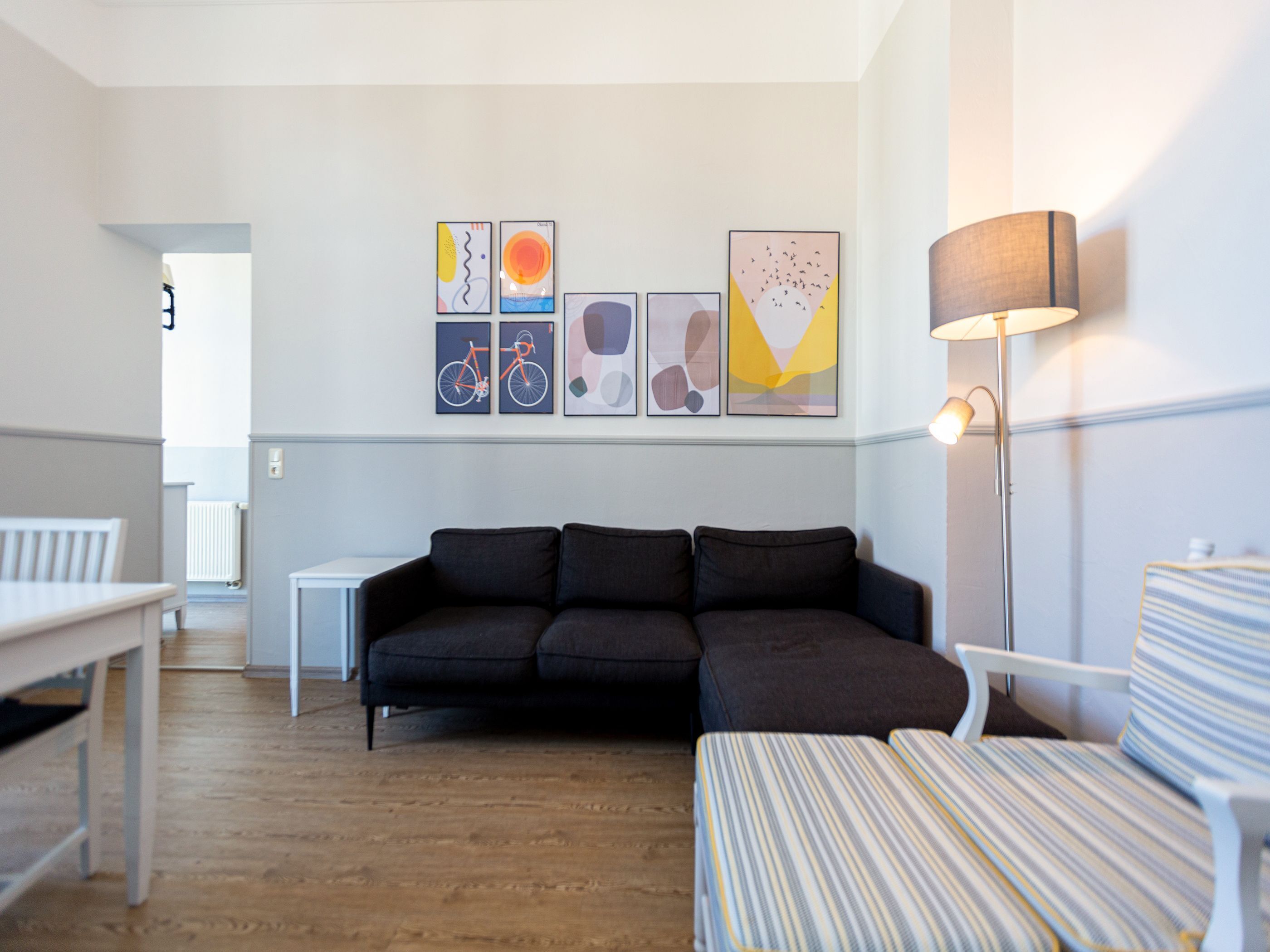Uma sala de estar moderna com um sofá escuro, poltronas listradas e um abajur. Há quadros abstratos na parede e uma mesa branca no canto.