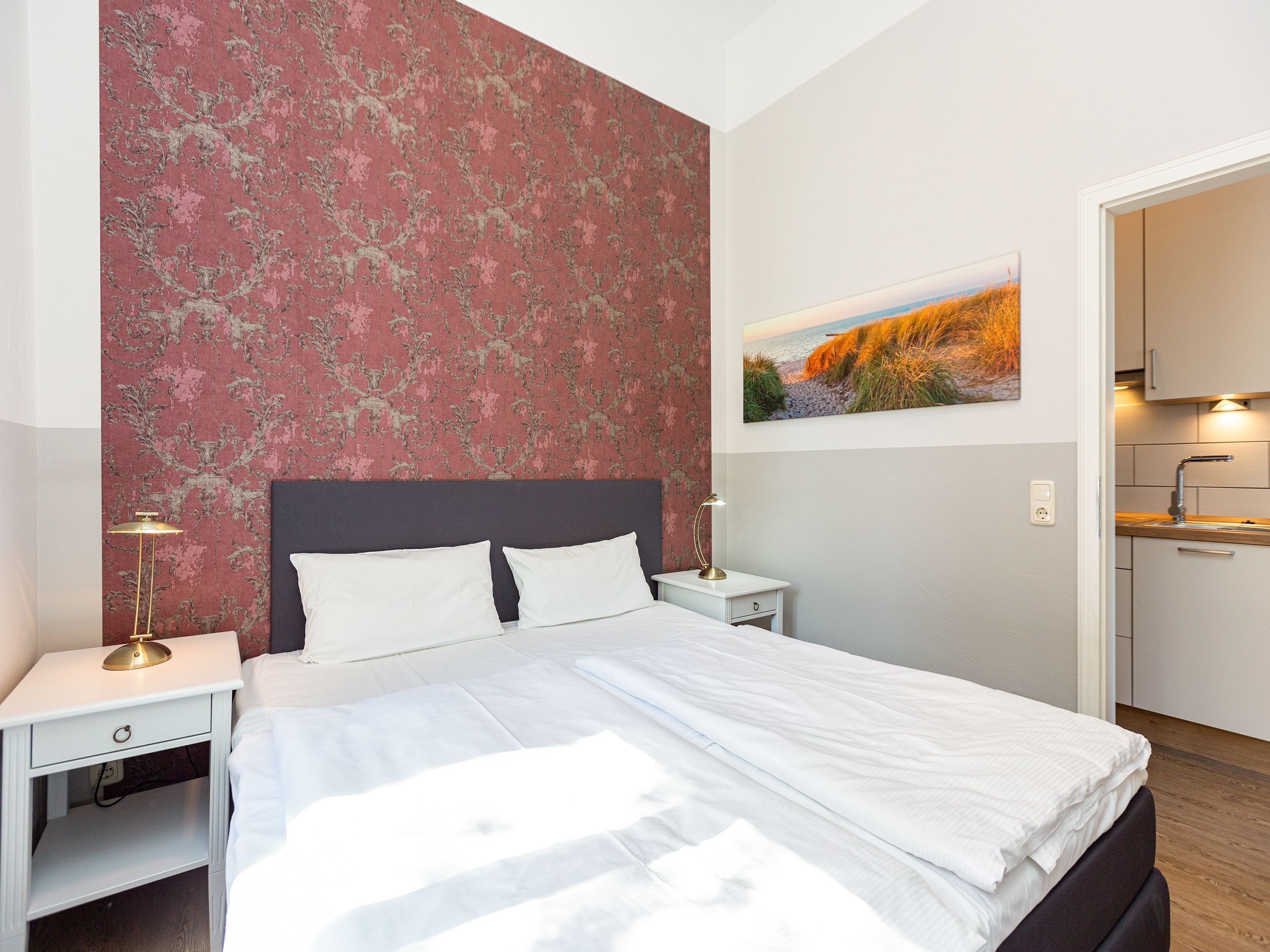 Een netjes opgemaakt bed met witte lakens, een nachtkastje met een lamp, een muur met een bloemenpatroon, en een schilderij van een strandgezicht.