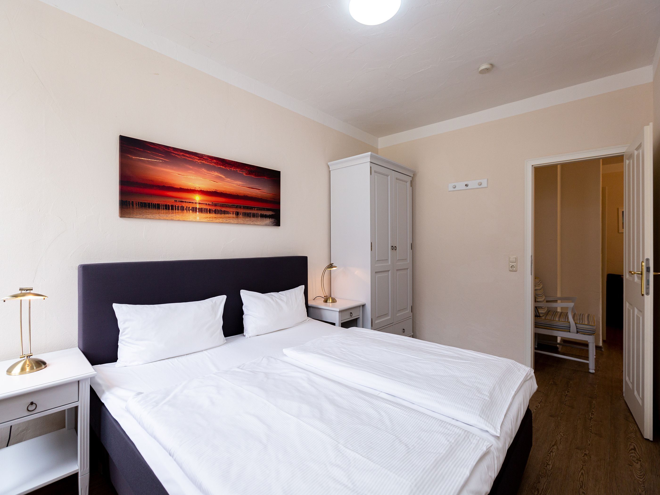Une chambre bien rangée, avec un grand lit, une table de chevet, une lampe, une armoire et un tableau accroché au mur.