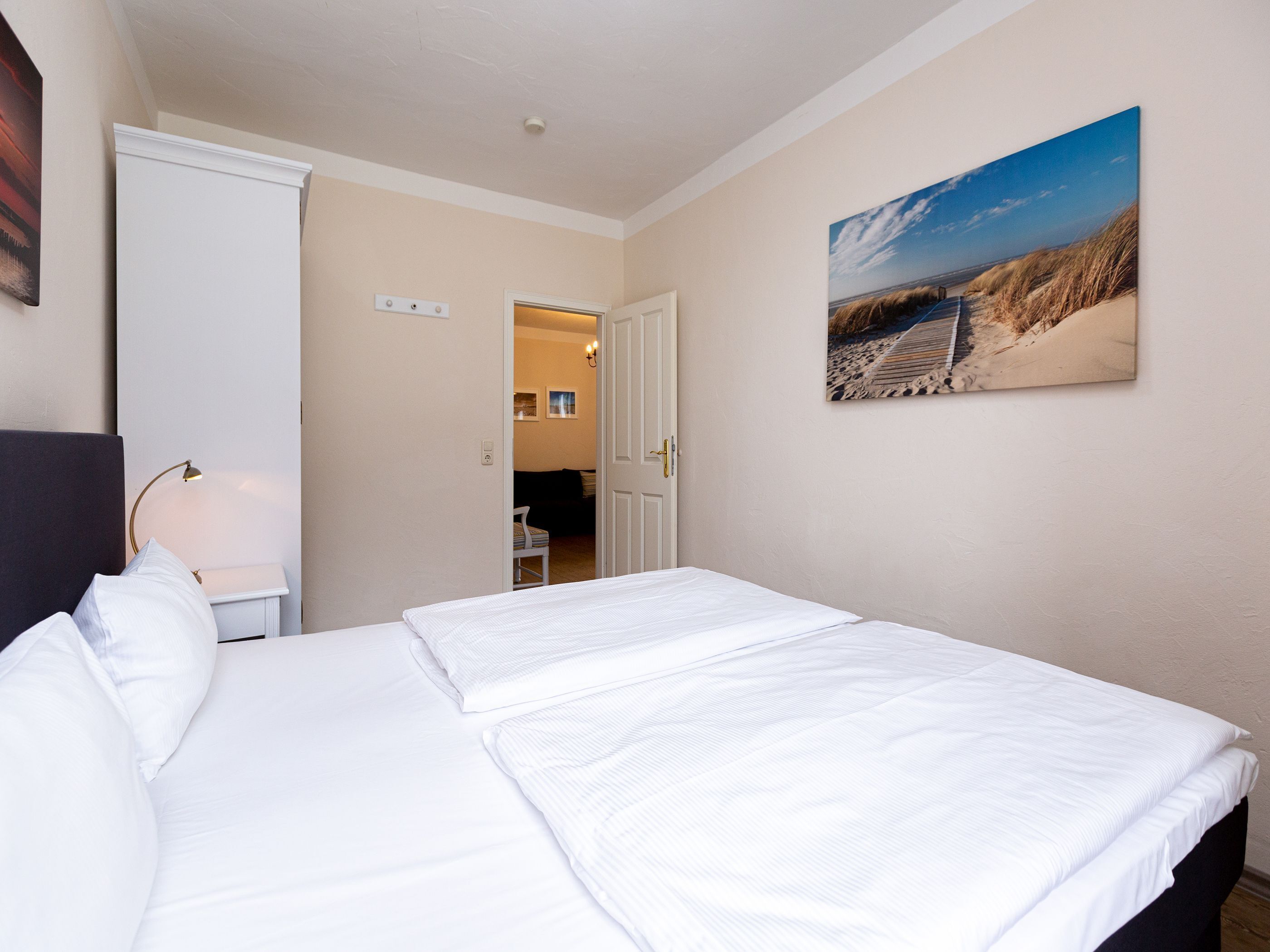 Ein ordentlich bezogenes Bett mit weißen Leinen, eine Nachttischlampe und ein Gemälde mit einer Strandansicht an der Wand.