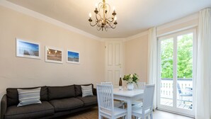 A room with a dark gray sofa, white chairs, a white table, and a chandelier. There are three framed pictures on the wall, and a window with curtains. - Villa Amelia, apt. 09 (Heringsdorf)