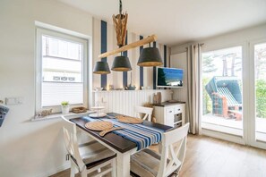 Ein Essbereich mit Holztisch, weißen Stühlen und einer Tischläufer mit Streifen. An der Wand ist ein Fernseher und ein Fenster mit Jalousien.