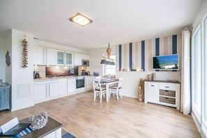 Eine moderne Küche mit weißen Schränken, einem Essbereich mit Holztisch und -stühlen sowie einem großen Flachbildfernseher an der Wand.