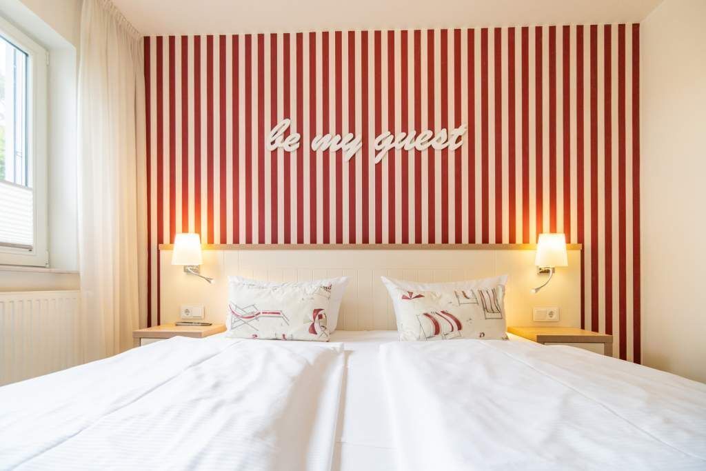 Ein Hotelzimmer mit einem Bett, zwei Nachttischlampen und einer Wand mit Streifen sowie einem Schild mit der Aufschrift 'be my guest'.