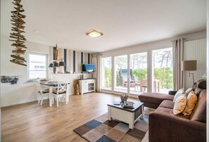 Ein modernes Wohnzimmer mit Sofa, Couchtisch und Essbereich. Ein Fernseher und ein Fenster mit Blick ins Grüne sind ebenfalls vorhanden.