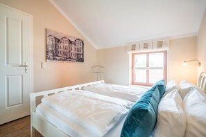Quarto com cama branca, janela com vista, porta e luminária fixada na parede.
