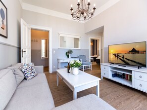 Un salon moderne avec un canapé, une table basse et un téléviseur. On y trouve un bureau avec une chaise et une bibliothèque. La pièce est dotée d’un plancher en bois et de murs blancs.