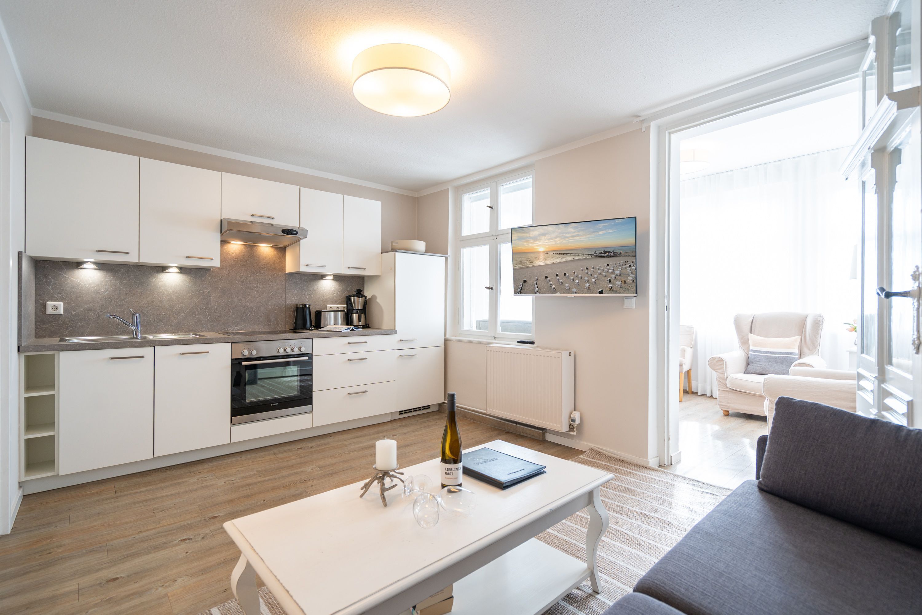 A modern kitchen with white cabinetry, a built-in oven, and a dining area with a white table and grey sofa.
