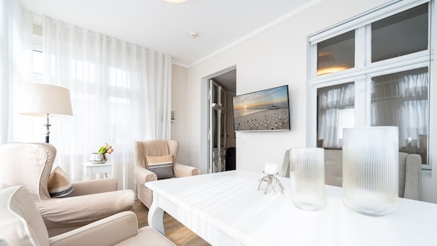 Quarto de hotel moderno com uma cama grande branca, uma escrivaninha, duas cadeiras, um abajur e uma pintura de uma paisagem praiana.
