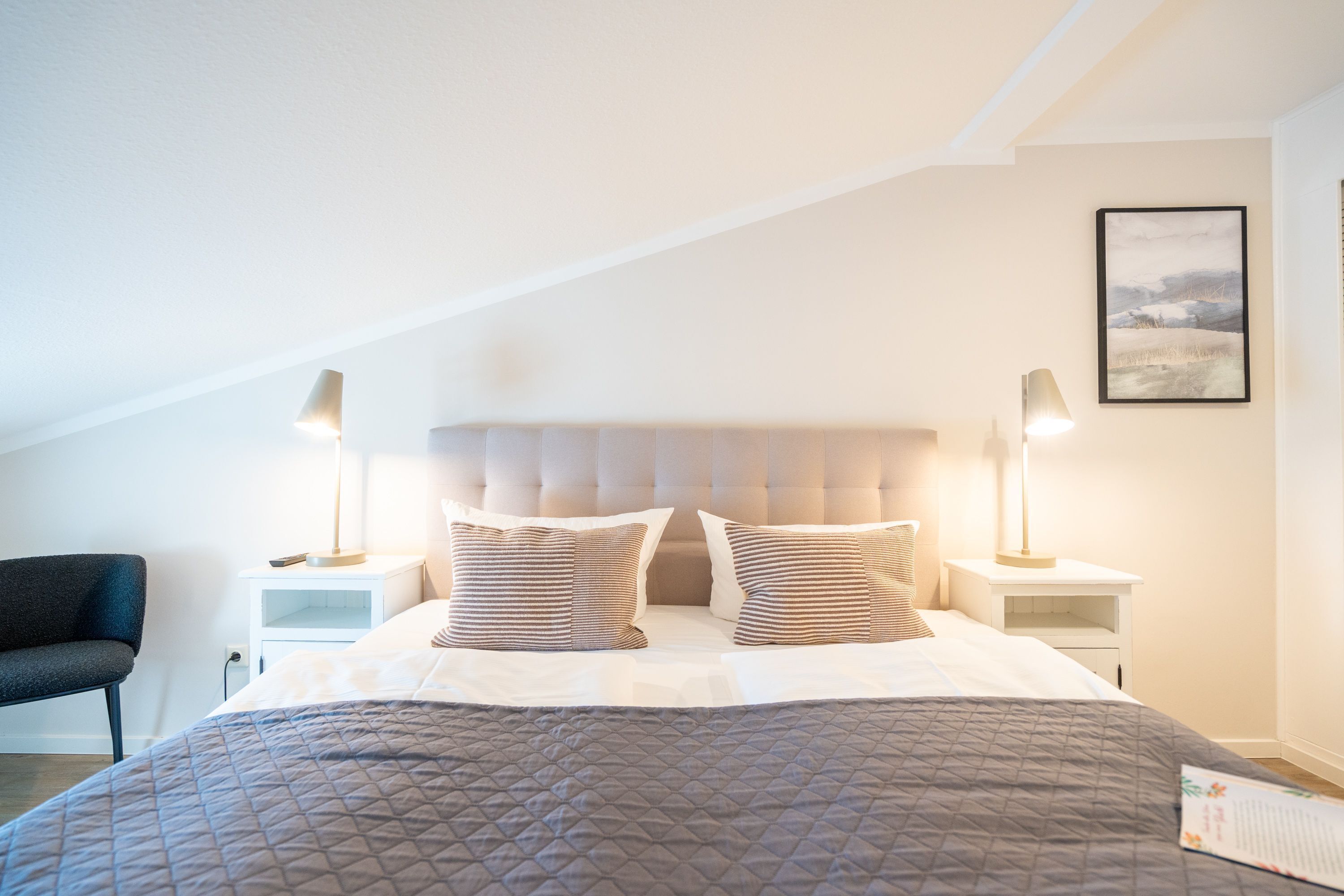 A bedroom with a large bed, two bedside tables, a chair, and a framed picture on the wall.