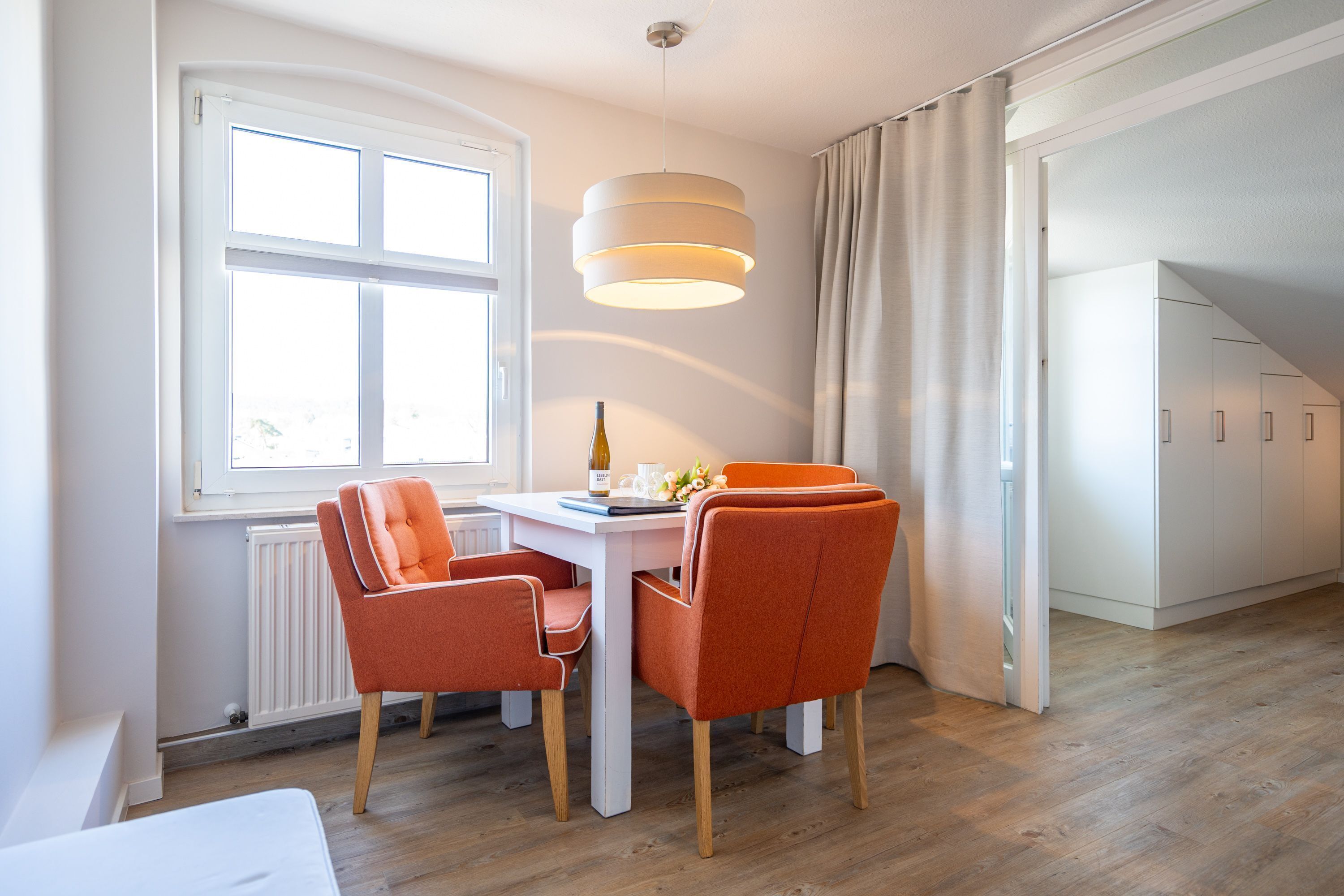 A modern dining area with a white table, orange chairs, a hanging light fixture, and a window with curtains.