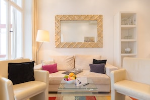 Een moderne woonkamer met een beige bank, twee fauteuils, een glazen salontafel met een fruitschaal, een lamp en een decoratieve spiegel.